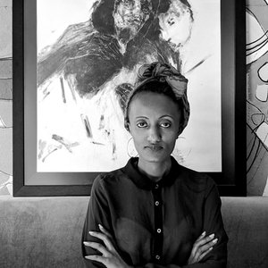 Photo en noir et blanc avec Tiemar Tegene en premier plan les bras croisé avec derrière elle une de ses oeuvres