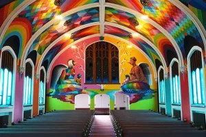Intérieur d'une église. Les murs et plafonds sont arc-en-ciel et sur le mur en face de nous se trouve 2 dessins d'animaux humanoïdes assis face à face